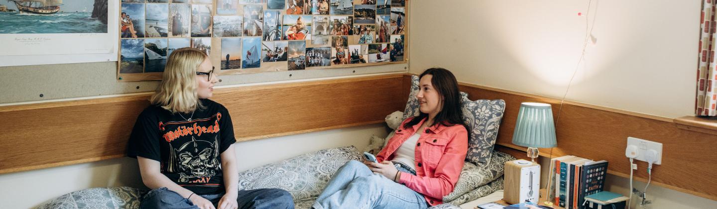 Two women students, one in a pink jacket and jeans and one in a black t shirt and jeans, sitting talking on a bed in a student room at Murray Edwards College, Cambridge.