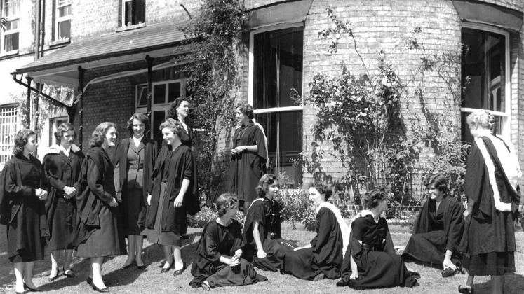 Black and white photo of a group of women in matriculation gowns, gathered on a lawn outside an elegant College building
