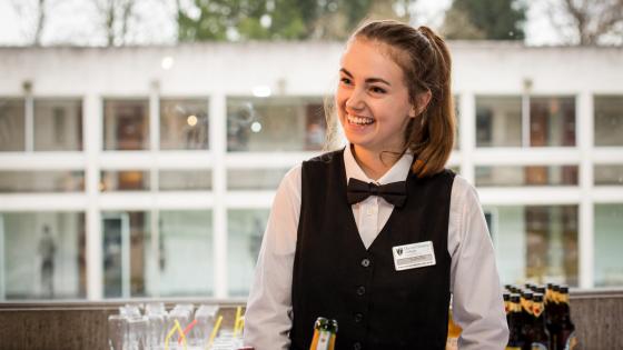 Waitress in white shirt with black waistcoat and bow tie serving Champagne and other drinks at an event at Murray Edwards College, Cambridge