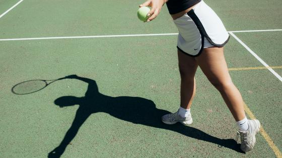 The shadow of a student holding a tennis raquet can be seen on the ground of the tennis court