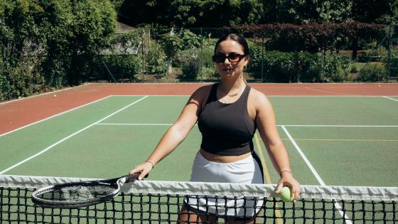 A student poses leaning on the net of the tennis court