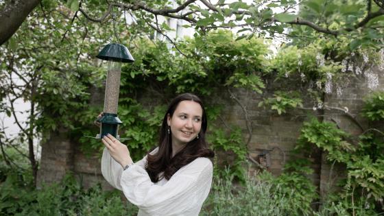 Young woman student in white top in gardens smiling and holding a bird feeder