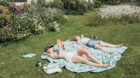 Two young women reading and sunbathing on a rug on a lawn by a flowerbed