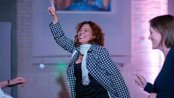 Woman with curly hair smiling and dancing in the middle of a dancefloor, one arm raised up pointing joyfully into the air.
