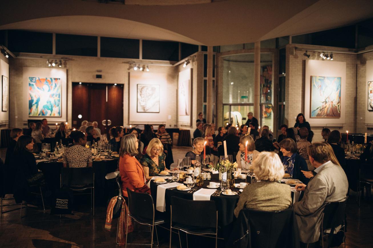 People sit and eat dinner by candlelight at several round tables. Artwork in the background.
