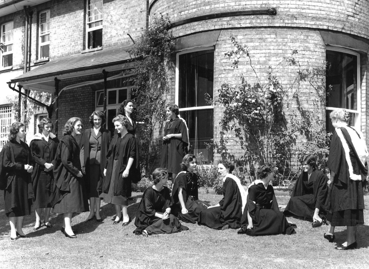 Black and white image of women students in gowns in front of an old brick house, 1954